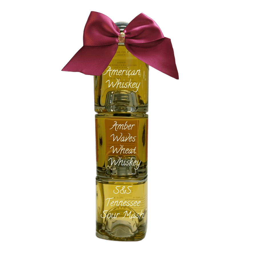 Three stacked glass bottles of whiskey with a large magenta satin bow tied around the neck, labeled 'American Whiskey', 'Amber Waves Wheat Whiskey', and 'S&S Tennessee Sour Mash' on a white background.