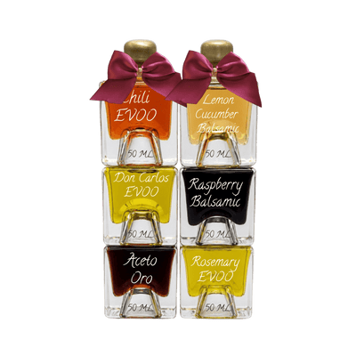 Six small glass bottles of EVOO balsams and accents arranged in a 2x3 grid on a white background. The bottles contain orange, yellow, black, and dark brown liquids, are topped with gold caps and purple bows, and feature white script labels reading 'Chili EVOO', 'Lemon Cucumber Balsamic', 'Don Carlos EVOO', 'Raspberry Balsamic', 'Aceto Oro', and 'Rosemary EVOO'.