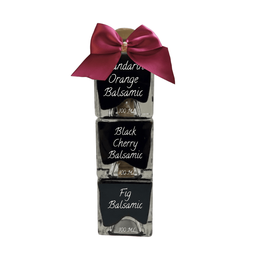 Three stacked glass bottles of balsamic syrup with black labels, tied together with a large magenta satin bow on top, shown against a plain white background.