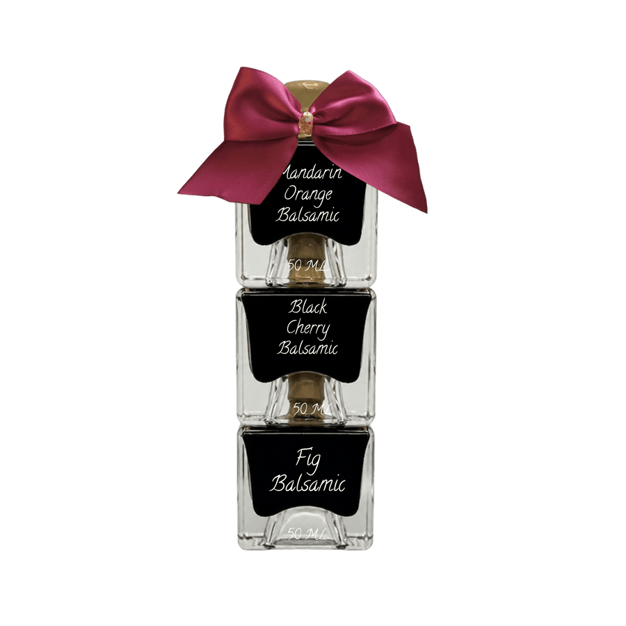 Three stacked clear glass bottles of scented hair oil with black labels reading 'Amandarin Orange Balsamic,' 'Black Cherry Balsamic,' and 'Fig Balsamic,' tied together with a large magenta satin bow on a white background.