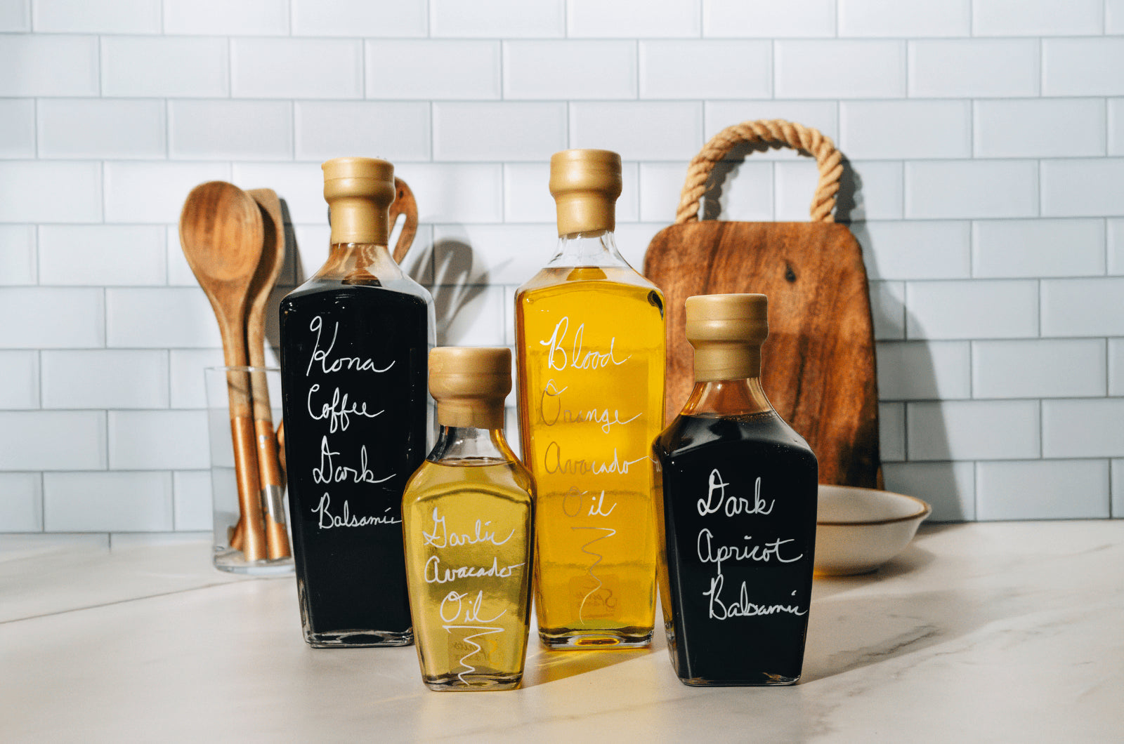 Five bottles of different types of oils on a kitchen counter with a tiled wall background.