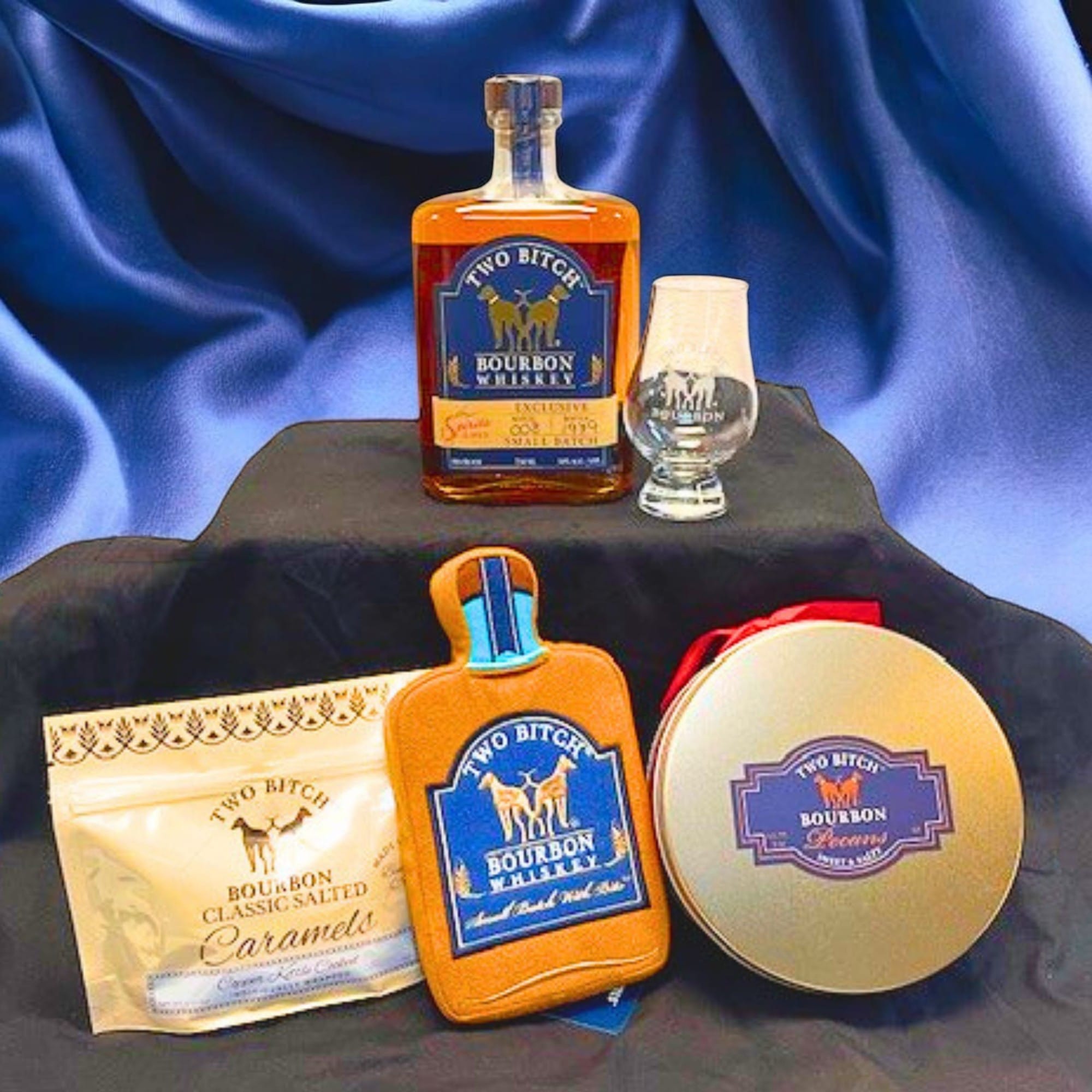 Collection of 'Two Bitch' Bourbon products including a bottle, glass, caramels, and a tin on a blue fabric background. Two bitch bourbon guft set