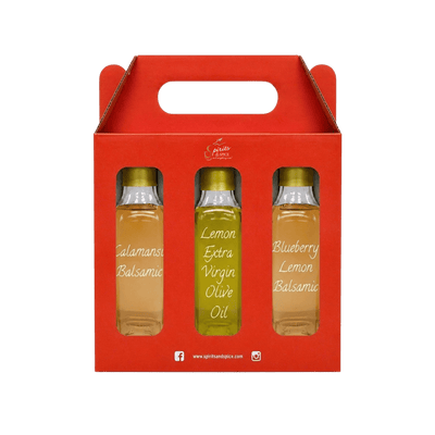 A red gift box with a handle containing three small glass bottles of oil. The center bottle is labeled 'Lemon Extra Virgin Olive Oil' and the side bottles are labeled 'Calamansi Balsamic' and 'Blueberry Lemon Balsamic'.