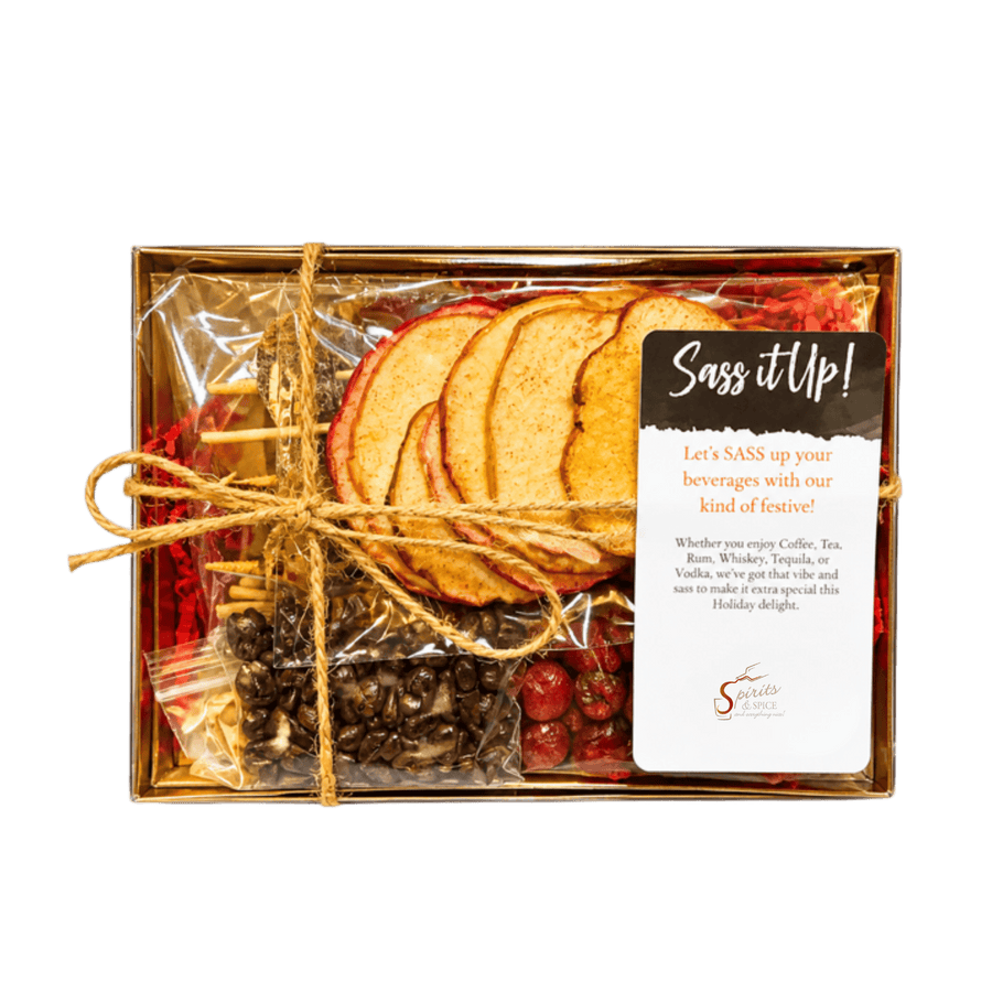 A boxed gift set from Sass itUp! featuring dried apple slices, red cranberries, coffee beans, tea bags, and holiday decorations inside a clear plastic-wrapped gold box with twine and a printed card reading 'Sass itUp!'.
