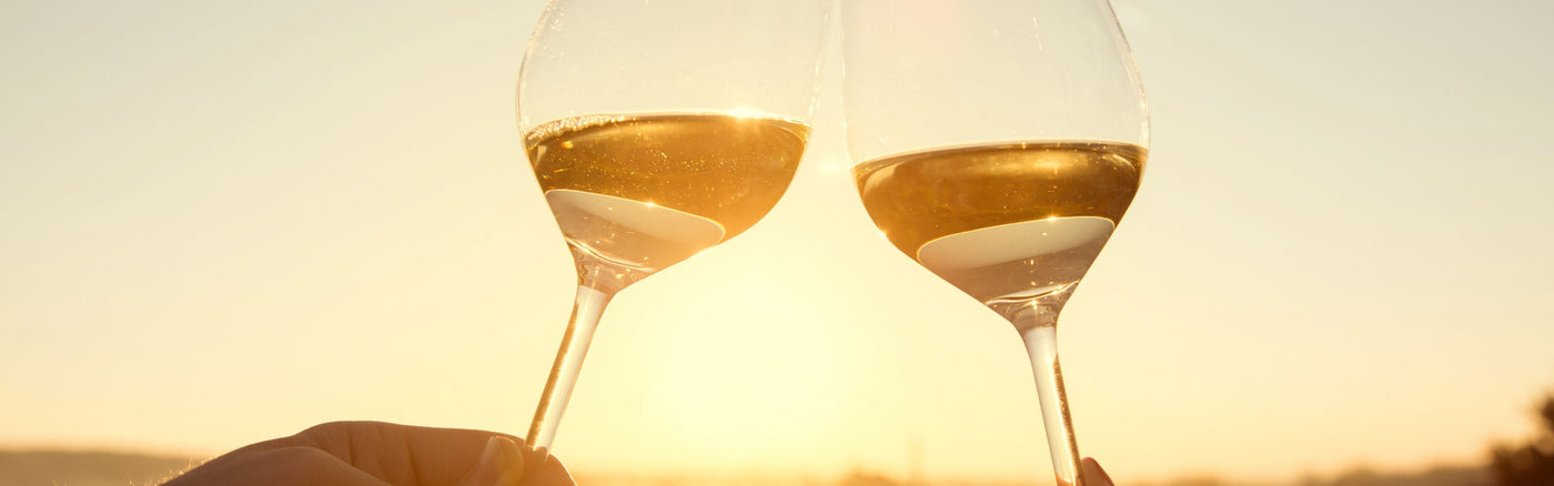 Two wine glasses filled with white wine clinking together in a toast, with a hand holding the glasses against a warm sunset sky.